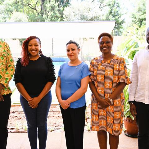 Five people stand side by side outside under cover, wearing a mix of patterned and solid-colour clothing and smiling at the camera.