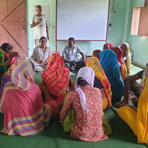 Group of women in colourful clothing seated in a circle indoors, listening to two facilitators, with a whiteboard behind them. 