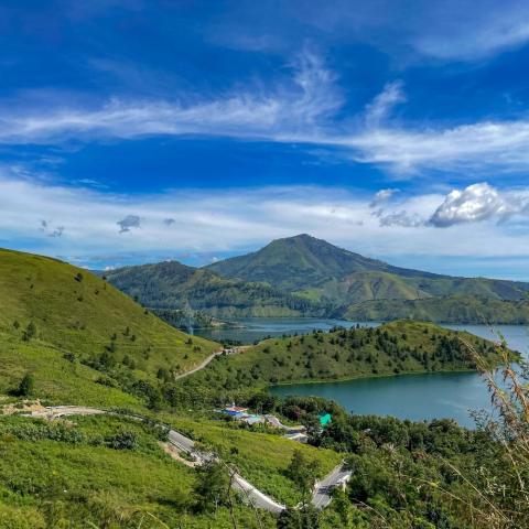 Green hills and a lake surrounded by mountains under a bright blue sky.