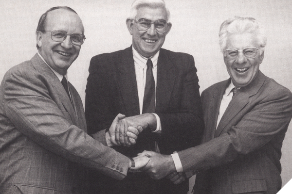 Black-and-white photo of three men in suits and ties all shaking each other’s hands and smiling happily. 