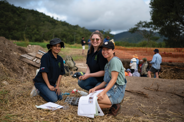 Three young women kneeling on the ground. One is holding a notebook. There are some digging tools, a spray bottle and backpacks on the ground. A group of people can be seen in a pit behind them. There are mountains in the background. 