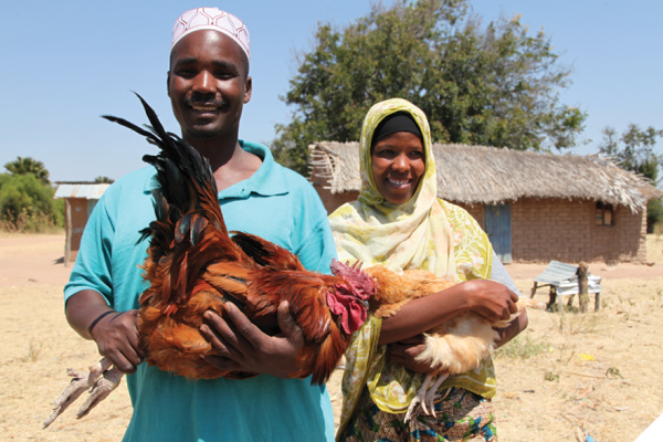 A man and a woman stand holding chickens in front of a mud hut with a thatch roof. 