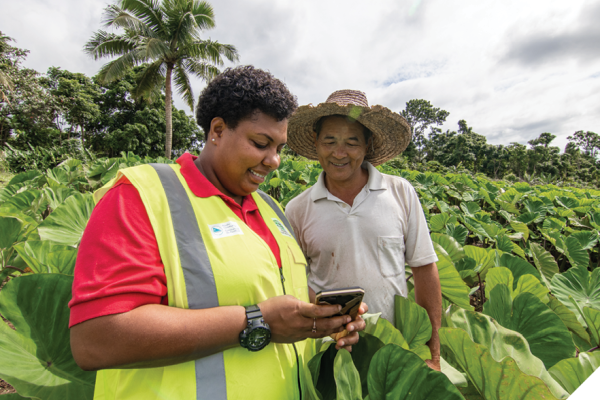 A man and a woman stand in a field of plants. The man is wearing a white polo shirt and a straw sun hat. The woman is wearing a red shirt with a bright yellow safety vest over top. They are both looking at an electronic device in the woman’s hands. 