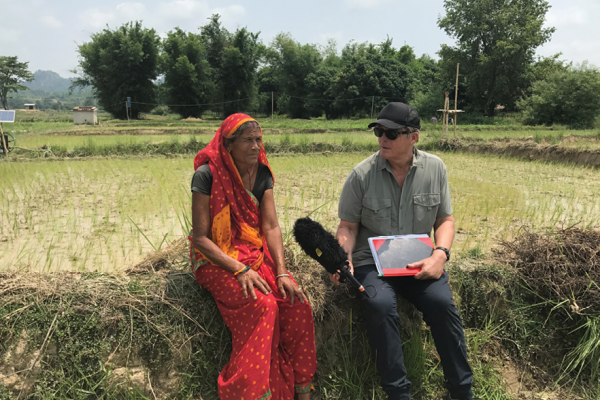 A man with a fuzzy microphone and a black cap sits next to a woman wearing a bright red dress and a red head scarf. They are sitting on the edge of a rice patty. He is interviewing her. 