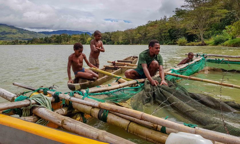 A floating fish farm in Yonki Reservoir in the Eastern Highlands Province of PNG, showing lush vegetation against high mountains in the background. A man and two young boys on a raft, pull a fish net.