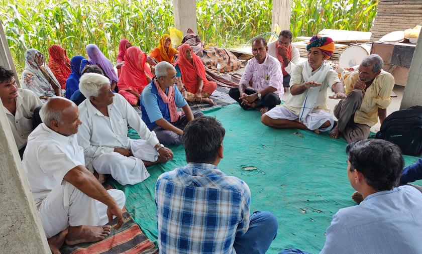 A group of adults sitting on the floor in a semi-circle under a shelter, engaging in a discussion. Some are wearing traditional Indian attire.