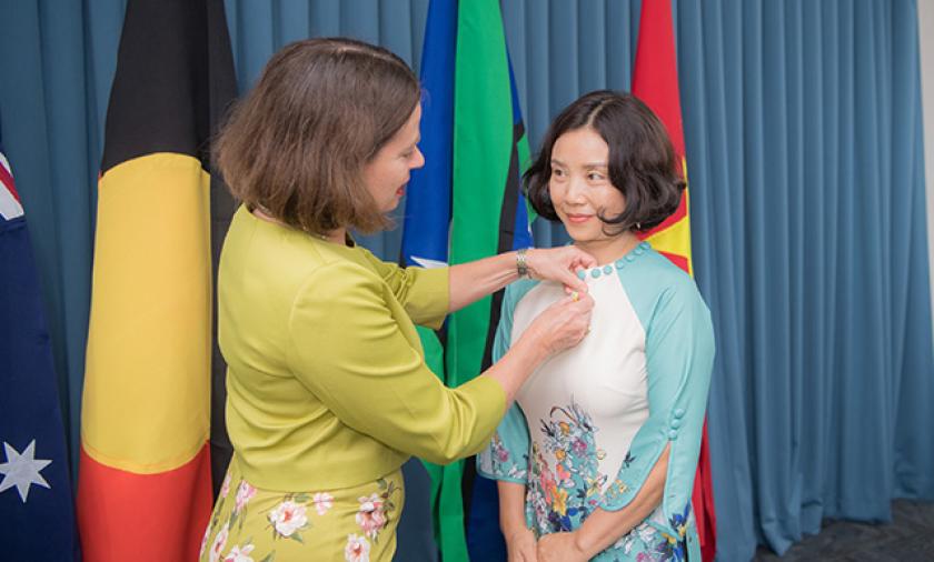One person pins a badge onto another in front of several flags.