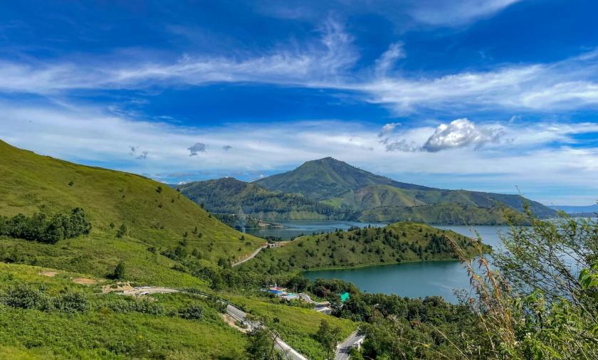 Green hills and a lake surrounded by mountains under a bright blue sky.